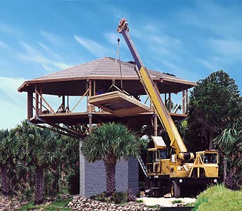 Elevated Hurricane-Resistant Oceanfront Pedestal Home Construction: 20 Ft Above Ground with Crane Installing Roof for Storm Surge Safety Crane lifting roof onto elevated 20-ft high oceanfront pedestal beach house during construction, hurricane-resistant design with palm trees and sandy site for storm surge and flood protection.